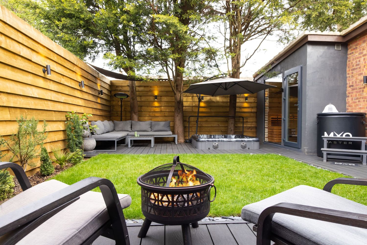 Garden spa patio in Bedfordshire with fire pit, outdoor seating, lounge chairs and landscaped lawn beside garden room.