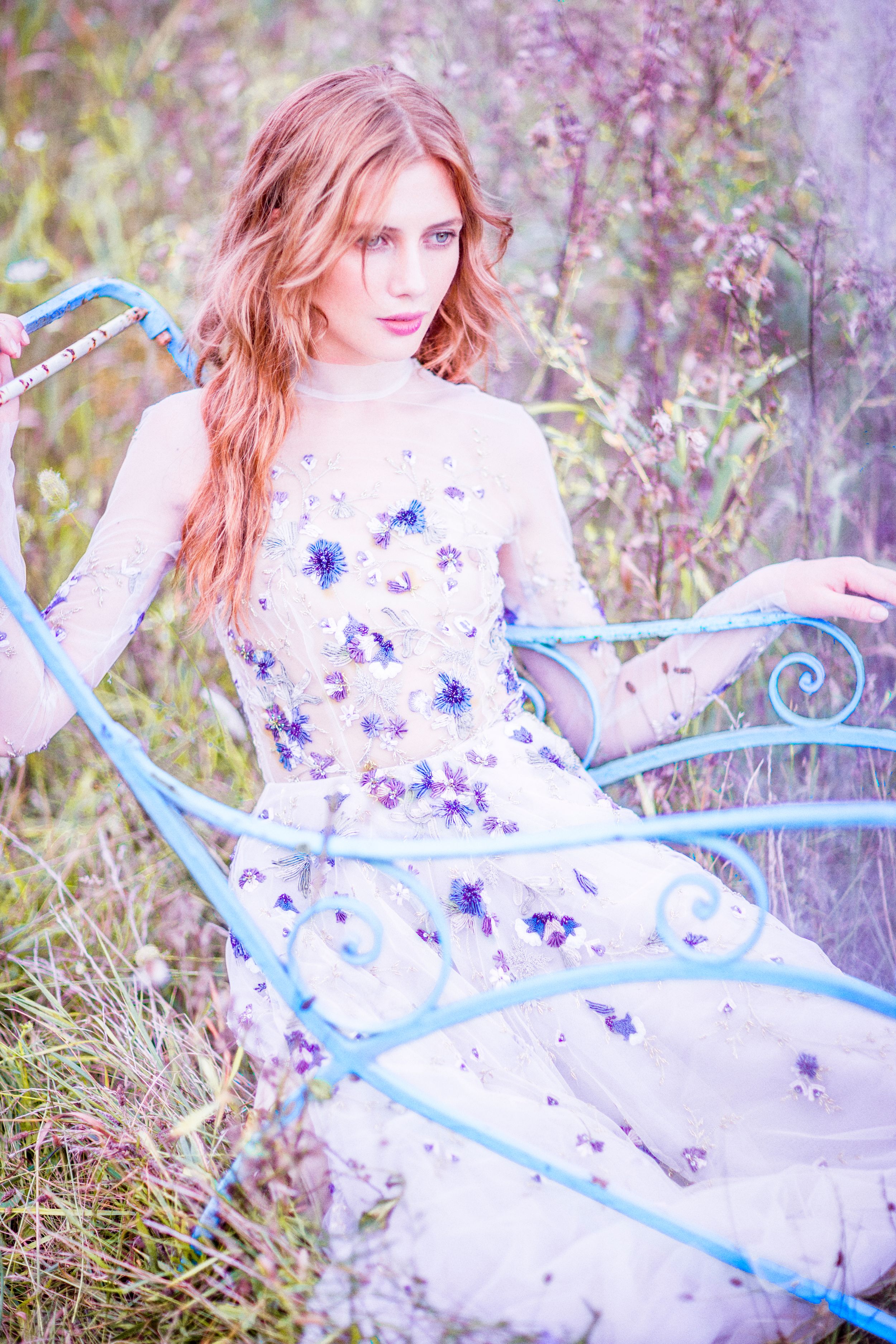 Young woman in floral white gown sitting in blue iron bed frame surrounded by wildflowers in dreamy outdoor setting.