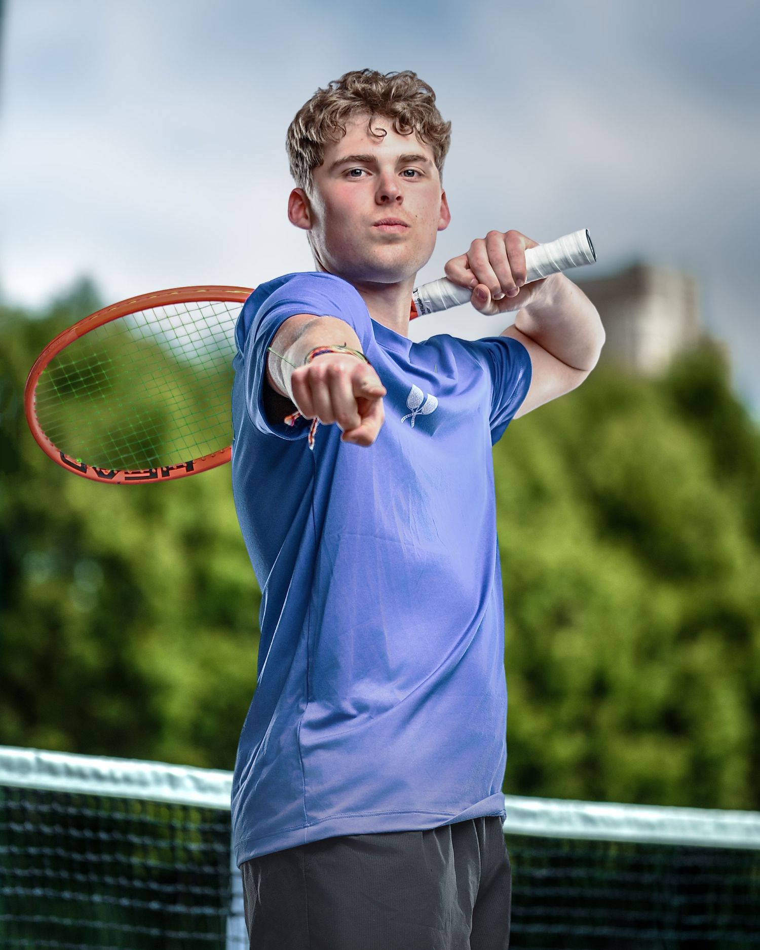 Young male tennis player in blue shirt pointing forward while holding a racket on an outdoor court.