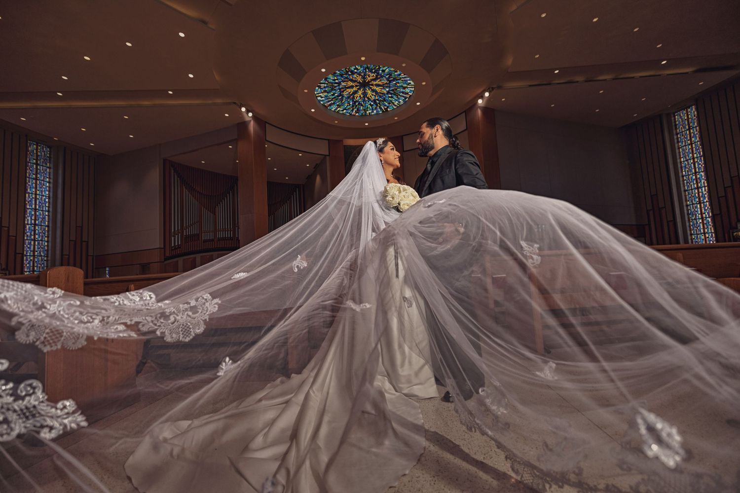 Dramatic bridal veil toss in a modern church under a stained glass skylight, featuring a lace-trimmed veil and white rose bouquet.