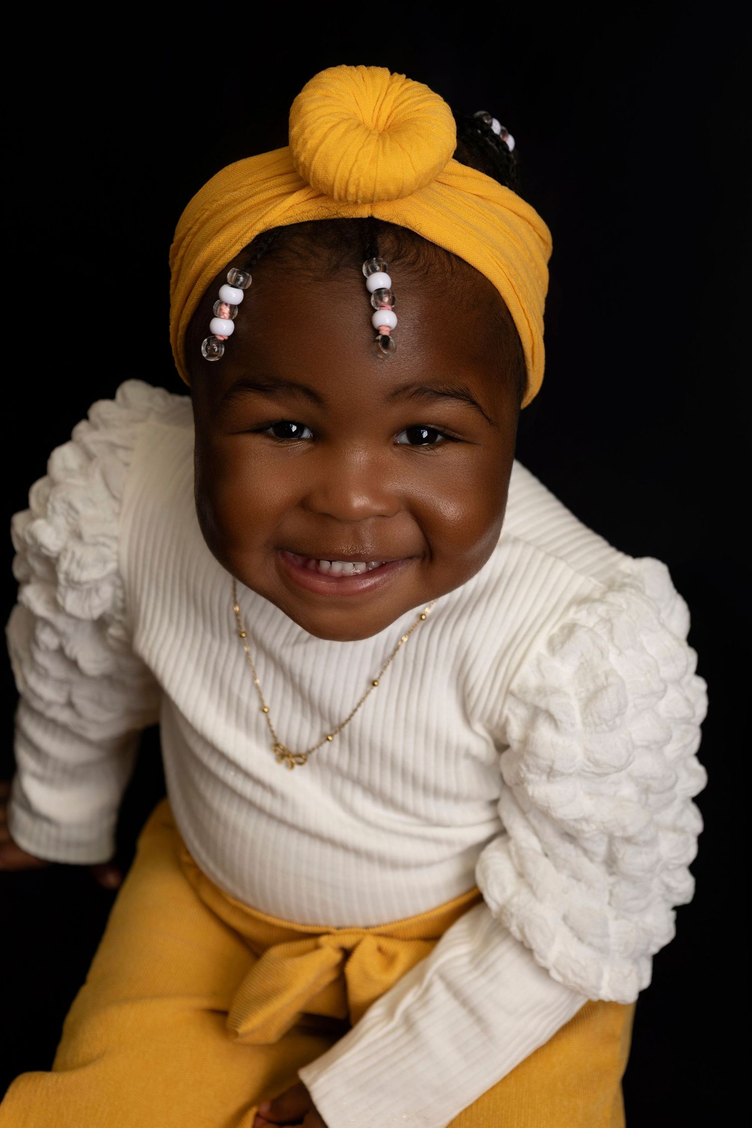 Smiling baby girl in yellow turban headband and white ruffled top with gold necklace on black background.