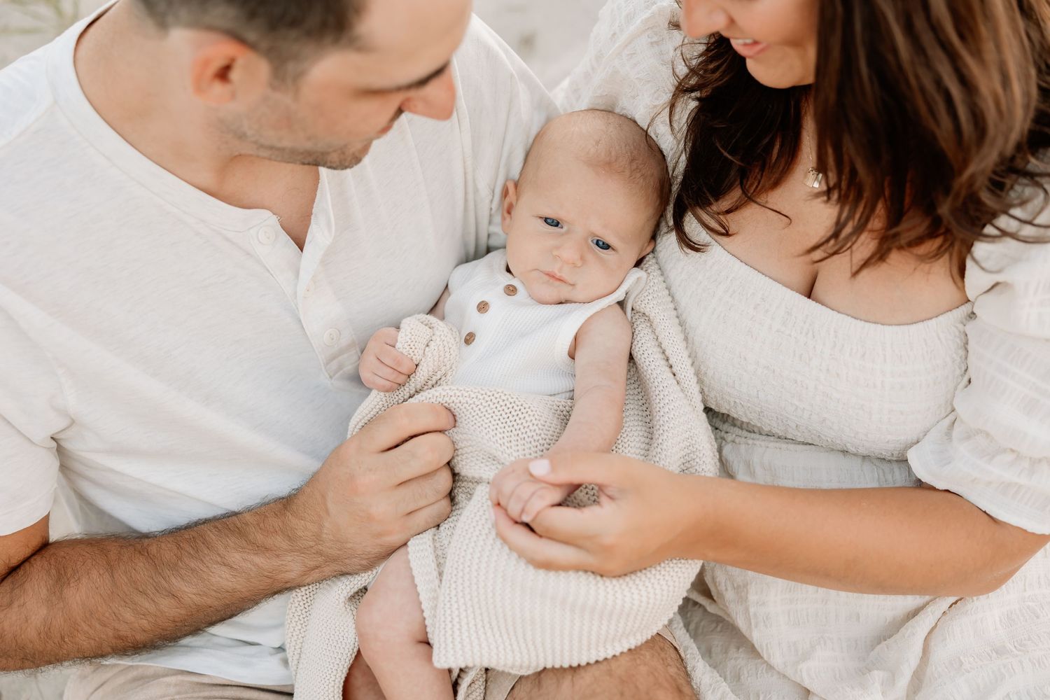 Parents lovingly gaze at their newborn baby wrapped in a soft cream blanket during a family portrait session.