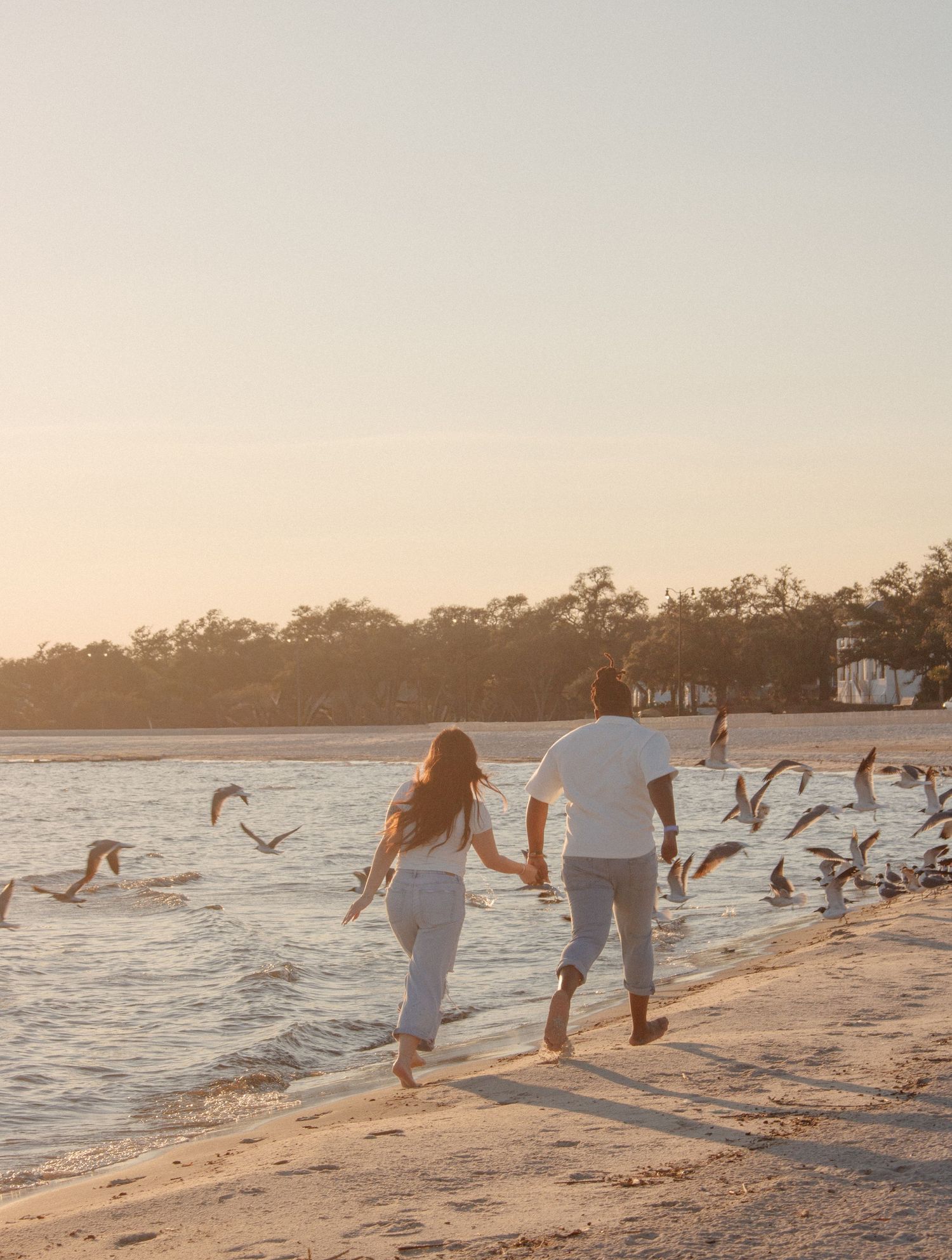A couple holds hands and runs along the shoreline at sunset as seagulls scatter over the shallow water.