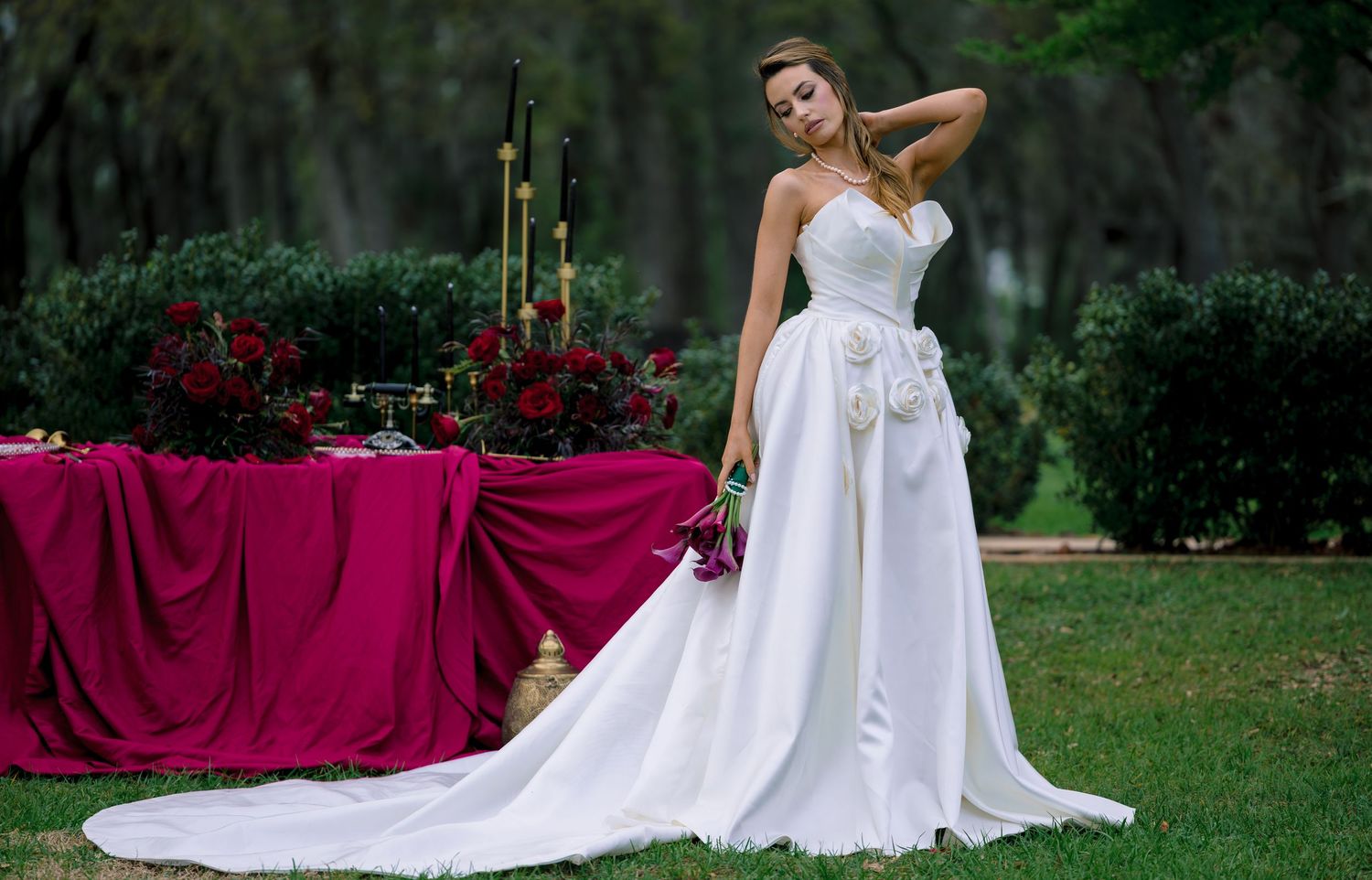 Elegant bride in white strapless gown with floral detail poses outdoors near a crimson-draped table with red roses.