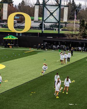 Oregon Ducks women's lacrosse team warming up on turf field with large yellow O logo sign in background.