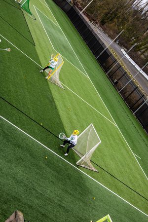 Aerial view of lacrosse players practicing on a green turf field with white lines and goals visible.
