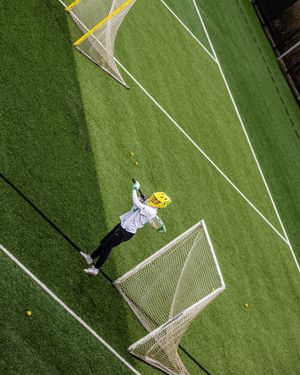 Aerial view of a lacrosse goalie in yellow helmet defending the net on a green turf field.