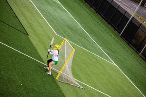 Aerial view of a lacrosse player in green and yellow helmet practicing near a goal on a turf field.