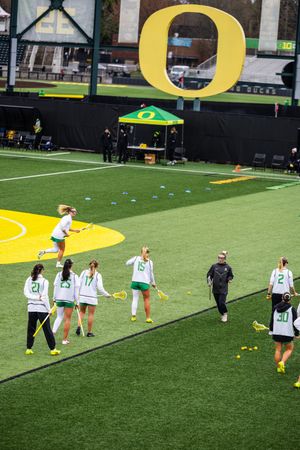 University of Oregon womens lacrosse team warms up on turf field beneath the iconic Oregon O logo.