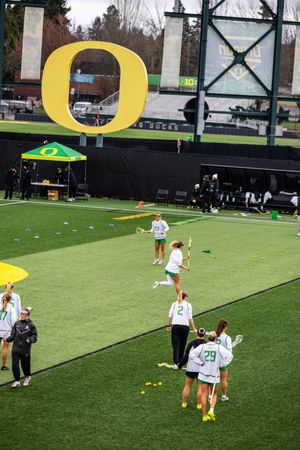 Oregon Ducks women's lacrosse team warming up on green turf field with large yellow O logo at Autzen Stadium.