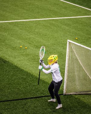 Female lacrosse player in yellow helmet holds stick near goal on green turf field during practice.