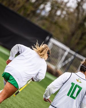 Oregon Ducks women's lacrosse player running on turf field during game, wearing white jersey and green shorts.