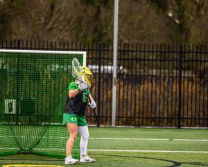 Oregon Ducks lacrosse goalie in green and yellow uniform stands ready in front of net on turf field.