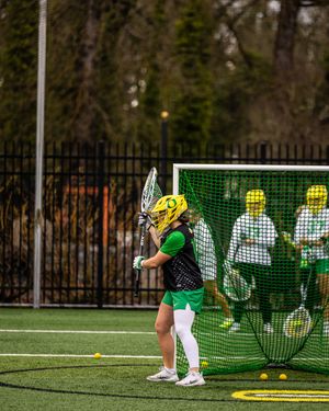 Female lacrosse player in green and yellow uniform practicing shots on goal during outdoor field training session.