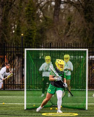Oregon Ducks lacrosse goalie in yellow helmet guards the net during a college game on a turf field.