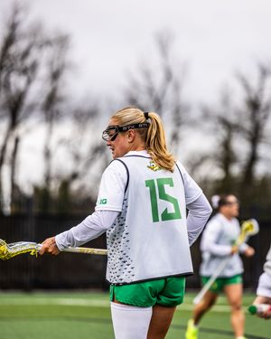 Oregon Ducks women's lacrosse player #15 in white and green uniform holding a lacrosse stick during an outdoor game.