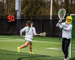 Two women's lacrosse players in white uniforms practice on a turf field, with player number 7 running toward the goal.