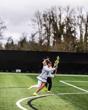 Female lacrosse player in white uniform cradles ball mid-game on green turf field with trees in background.