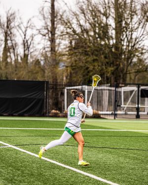 Female lacrosse player in white Oregon Ducks uniform winds up for a shot on an outdoor turf field.