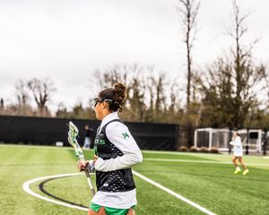 Female lacrosse player in green uniform holding a stick on an outdoor turf field during a cloudy day.