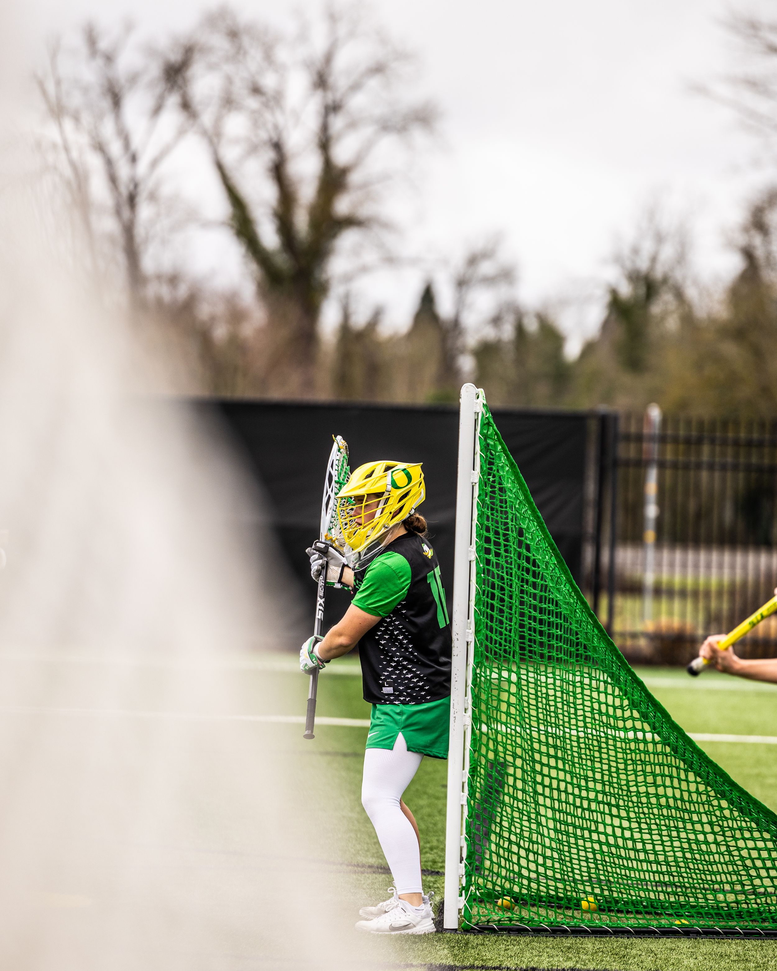 Lacrosse goalie in green and yellow gear stands ready at the net during an outdoor game.