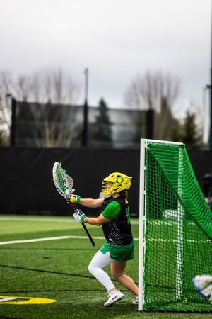 Oregon Ducks lacrosse goalie in green and yellow uniform defending the net during an outdoor game.