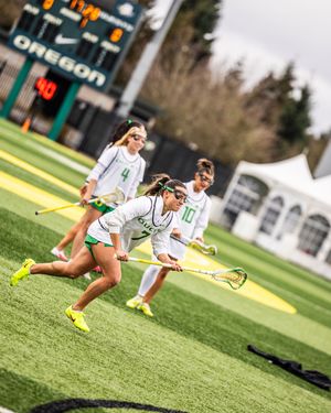Oregon Ducks women's lacrosse players in white uniforms sprint down turf field during competitive game near team scoreboard.
