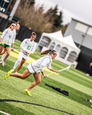 Oregon women's lacrosse player in white uniform sprints with stick during outdoor game on turf field.