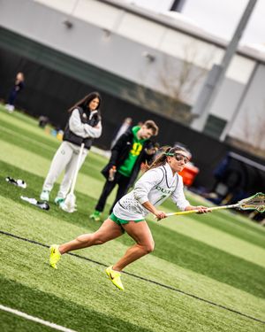 Female lacrosse player in white and green uniform sprints on turf field during practice, coaches watching in background.