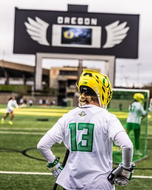 Oregon Ducks lacrosse player #13 in white jersey and yellow helmet stands on field with scoreboard behind.