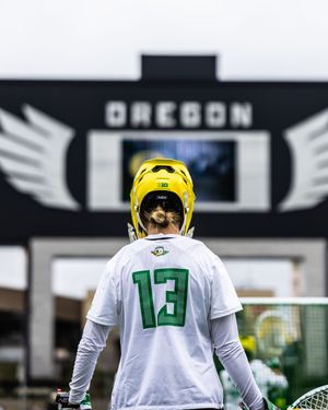 Oregon Ducks lacrosse player #13 in white jersey and yellow helmet stands before the Oregon scoreboard.