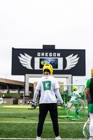 Oregon Ducks lacrosse player #13 stands on field facing scoreboard with iconic wing logo at Autzen Stadium.