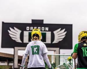 Oregon lacrosse player wearing number 13 jersey stands before large Oregon Wings logo sign.