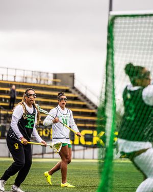 Oregon Ducks women's lacrosse players compete on field, wearing white and green uniforms with yellow cleats.