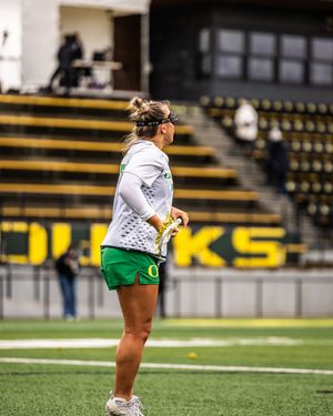 Female lacrosse player in Oregon Ducks green and white uniform holding a stick on the field.