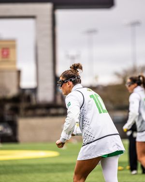 Female lacrosse player in white Oregon Ducks uniform number 10 prepares to throw during an outdoor collegiate game.