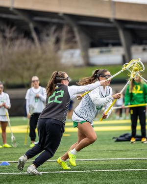 Two women's lacrosse players in green uniforms practicing stick skills on turf field.
