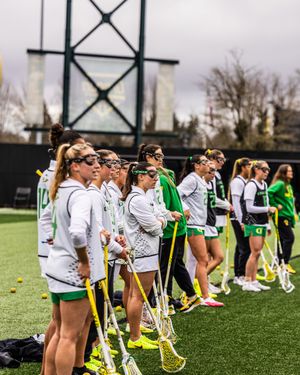 Oregon Ducks women's lacrosse team lined up on the sideline in white and green uniforms holding lacrosse sticks.