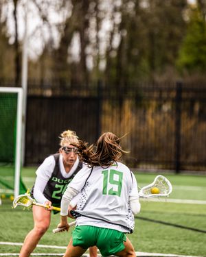 Two female lacrosse players in green and white Oregon Ducks uniforms compete on an outdoor field.