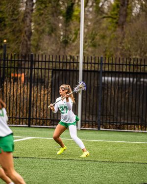 Female lacrosse player #27 in white and green uniform cradles ball on turf field during outdoor game.