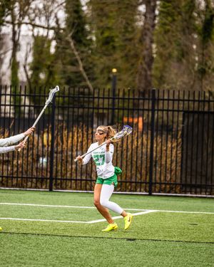 Female lacrosse player #27 in green uniform winds up for a shot during an outdoor game on turf field.