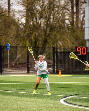 Female lacrosse player in green uniform holds stick on turf field during game with scoreboard showing 90.
