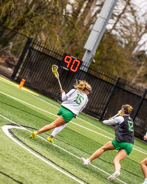 Female lacrosse player #15 in green and white uniform winds up to shoot on a turf field during a competitive game.