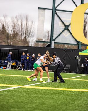 Oregon Ducks women's lacrosse player competing in a face-off with referee on green turf field.