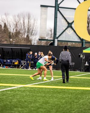 Two women's lacrosse players battle for possession on a green turf field as a referee watches nearby.