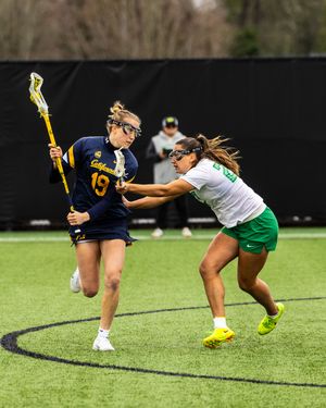 Female lacrosse player in navy jersey #19 dodges a defender in green during a competitive women's college lacrosse game.