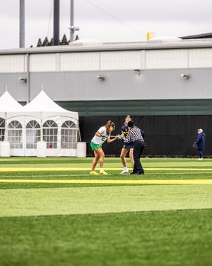 Two female lacrosse players face off on a green turf field with a referee nearby and white tents in the background.