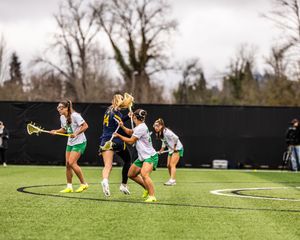 Women's lacrosse players compete for possession during a college game on a green turf field.