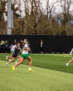 Women's lacrosse players compete on a turf field, with players in green and navy uniforms running during a game.