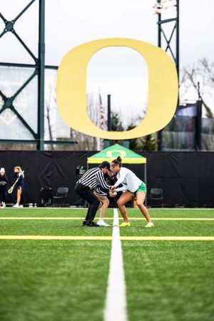 Oregon Ducks women's lacrosse player faces off with referee on turf field under large gold O logo.